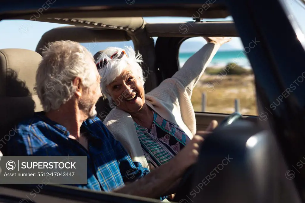 Side view close up of a senior Caucasian couple sitting in their car, driving to the beach in the sun, the man at the wheel and the woman with her hand out of the window and both smiling at each other