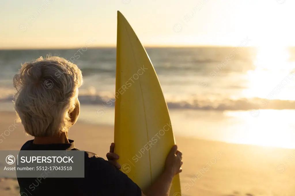 Rear view of a senior Caucasian woman at the beach at sunset, standing on the sand holding a surfboard and looking out to sea