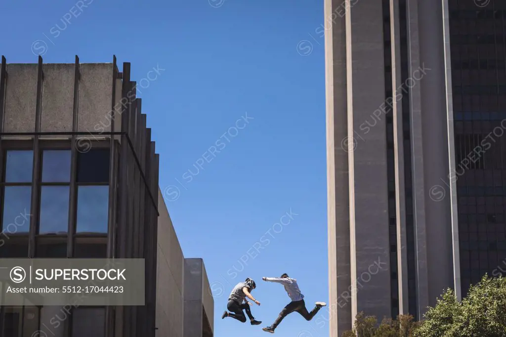 Side view of two Caucasian men practicing parkour by the building in a city on a sunny day, jumping up between modern buildings.