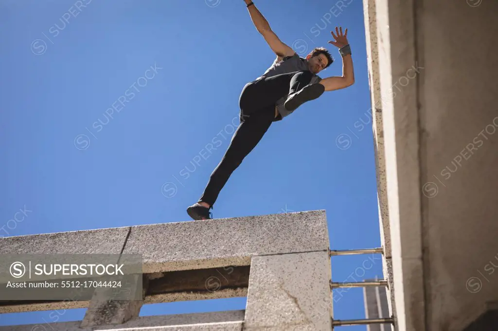 Front low angle view of a Caucasian man practicing parkour by the building in a city on a sunny day, jumping on the rooftop.