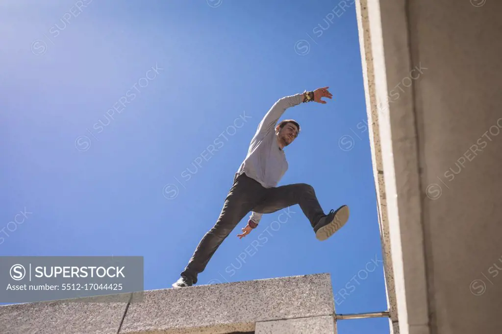 Front low angle view of a Caucasian man practicing parkour by the building in a city on a sunny day, jumping on the rooftop.