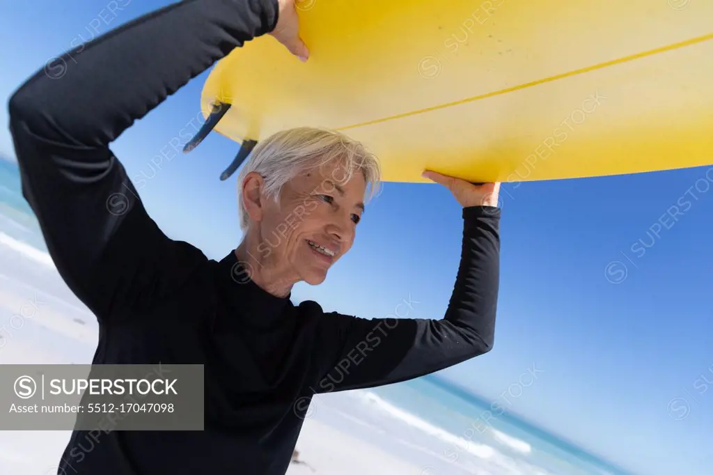 Senior Caucasian woman enjoying time at the beach, standing and holding a surfboard above her head