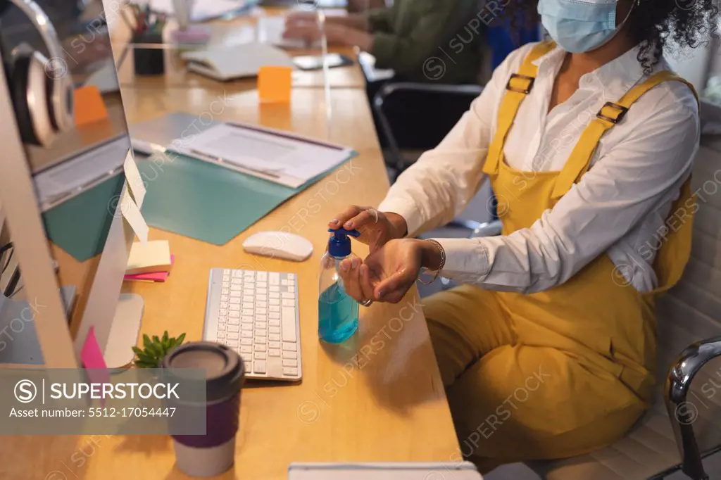 Mixed race female creative sitting at a desk in an office disinfecting hands with hand santiser. Health and hygiene in workplace during Coronavirus Covid 19 pandemic.