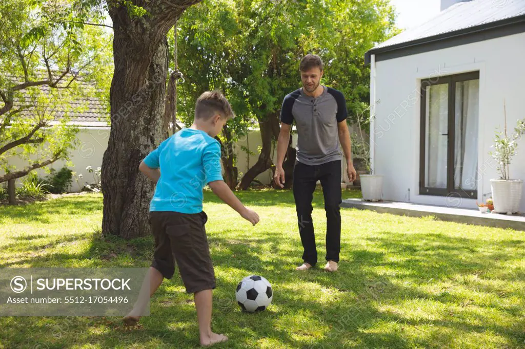 Caucasian man spending time with his son together, playing football in garden. Social distancing during Covid 19 Coronavirus quarantine lockdown.