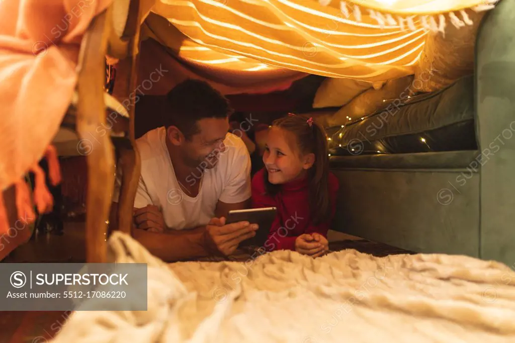 Caucasian man with his daughter smiling and using tablet, lying in blanket fort. quality lifestyle family time together.