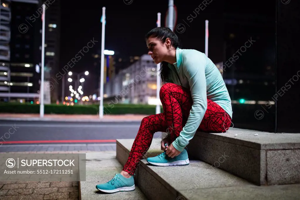 Side view of a fit Caucasian woman with long dark hair wearing sportswear exercising outdoors in the city in the evening, taking a break from her workout sitting on stair tying shoelaces with urban buildings in the background.