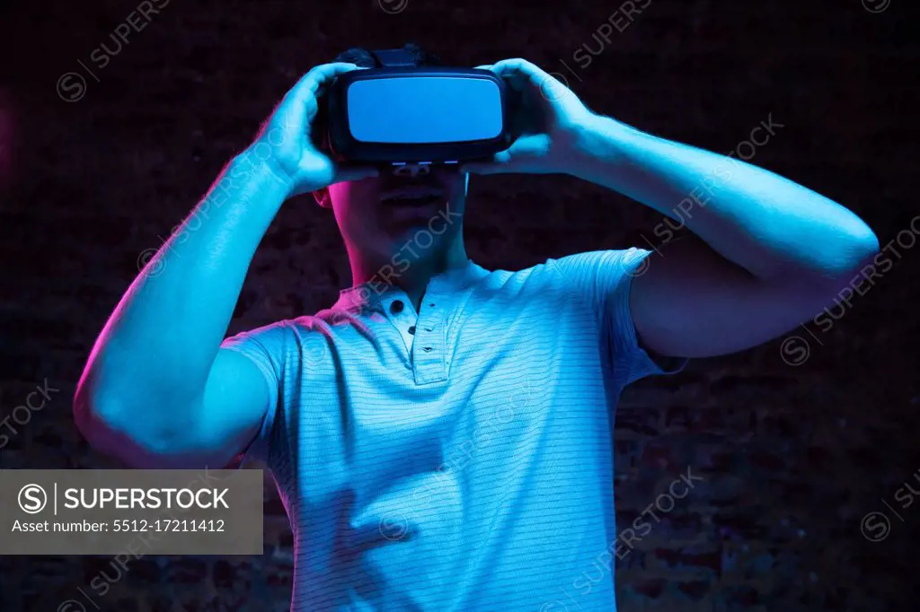Front view of a Caucasian man wearing a Virtual Reality headset and looking around, lit with pink and blue light on a black background.