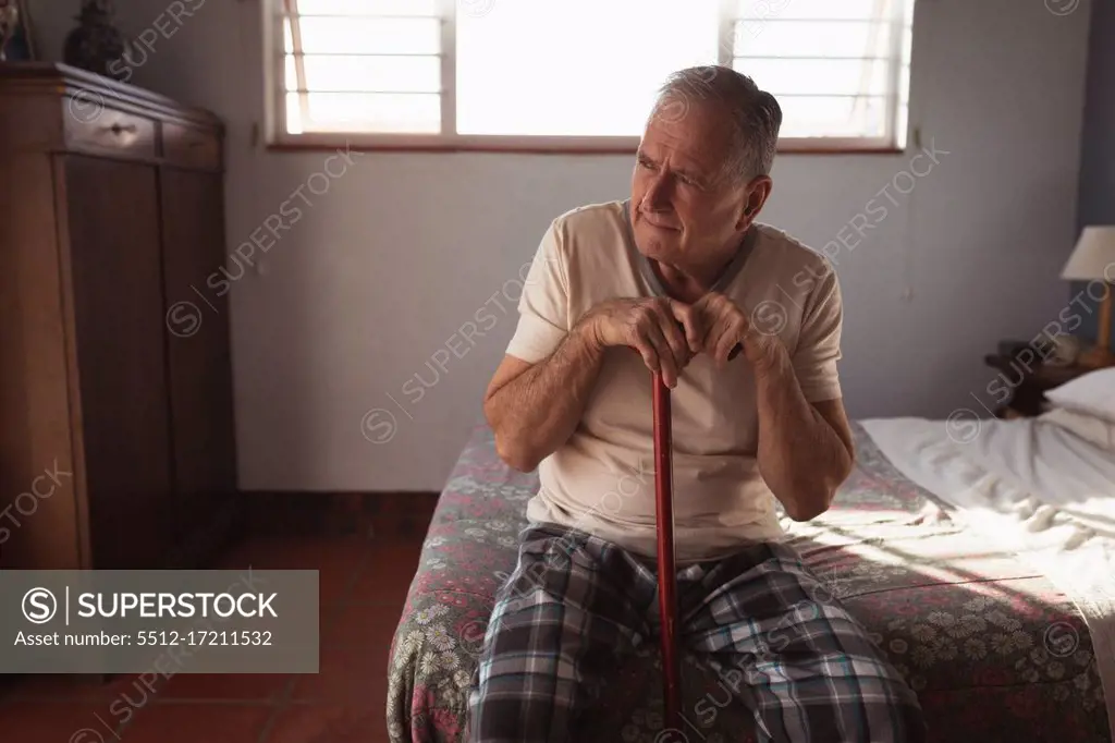 Front view of a senior Caucasian man relaxing at home in his bedroom, sitting on the side of the bed holding his cane and thinking after getting up in the morning