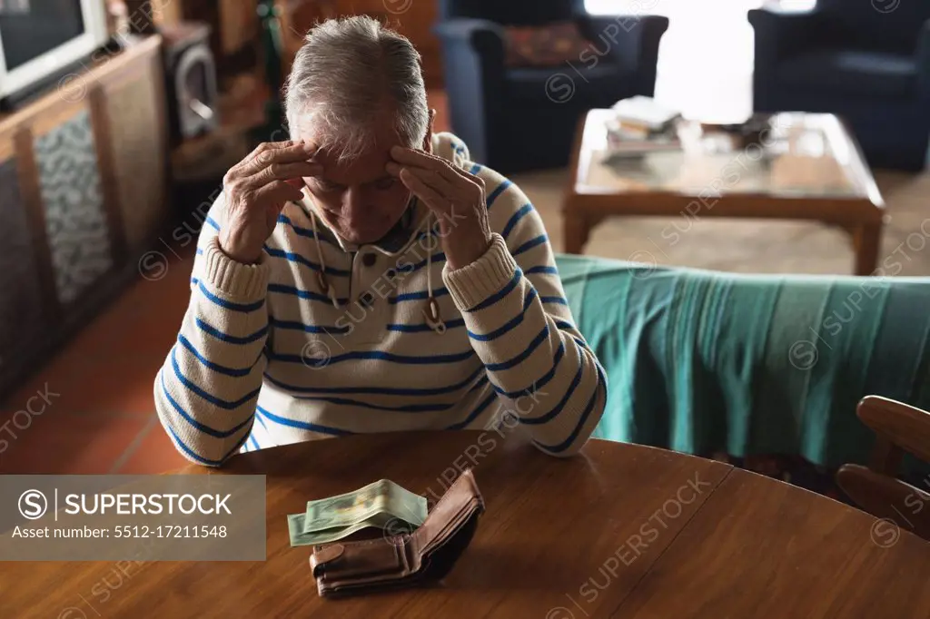 Front view of a senior Caucasian man at home, sitting at the table in his dining room, looking at the money in his wallet, holding his head and thinking