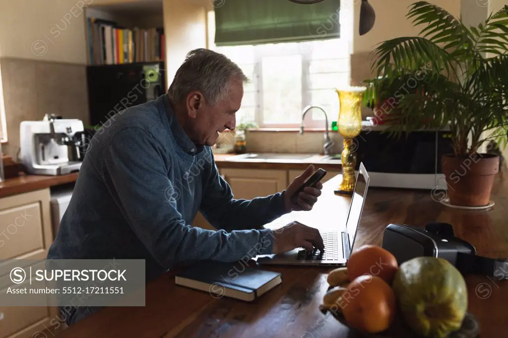 Side view of a senior Caucasian man relaxing at home, sitting at the counter in his kitchen using a smartphone and laptop computer