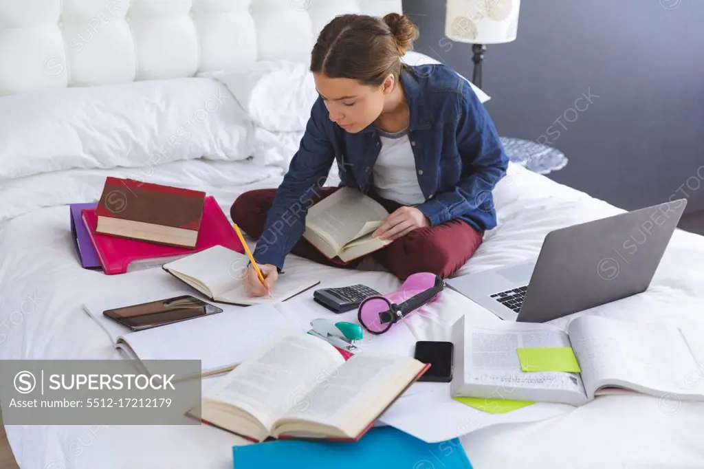 Caucasian woman spending time at home, sitting on bed in bedroom studying from home, holding book and making notes. Social distancing during Covid 19 Coronavirus quarantine lockdown.