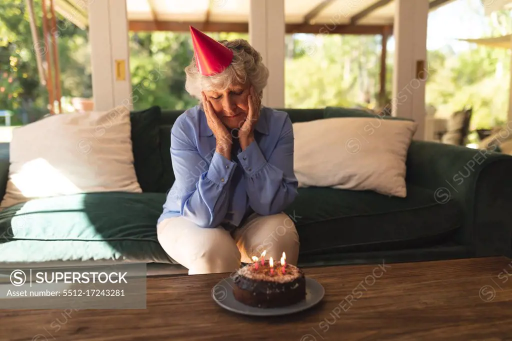 Senior caucasian woman spending time at home celebrating a birthday, wearing party hat and looking at cake. self isolation at home during coronavirus covid 19 quarantine lockdown.