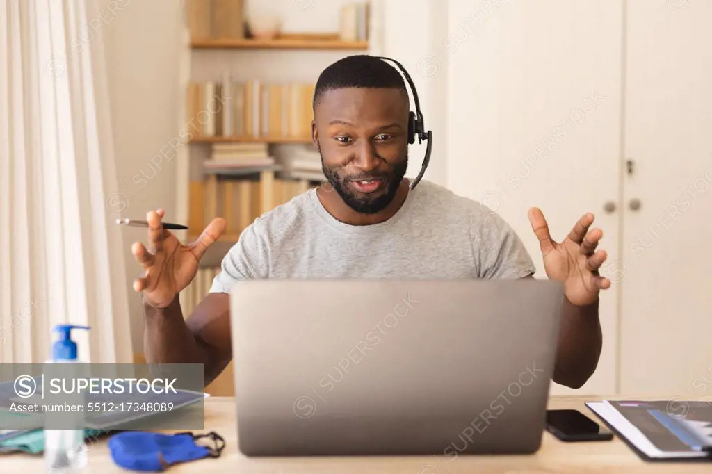 African american man using phone headset while having a video chat on laptop while working from home. social distancing during covid 19 coronavirus quarantine lockdown.