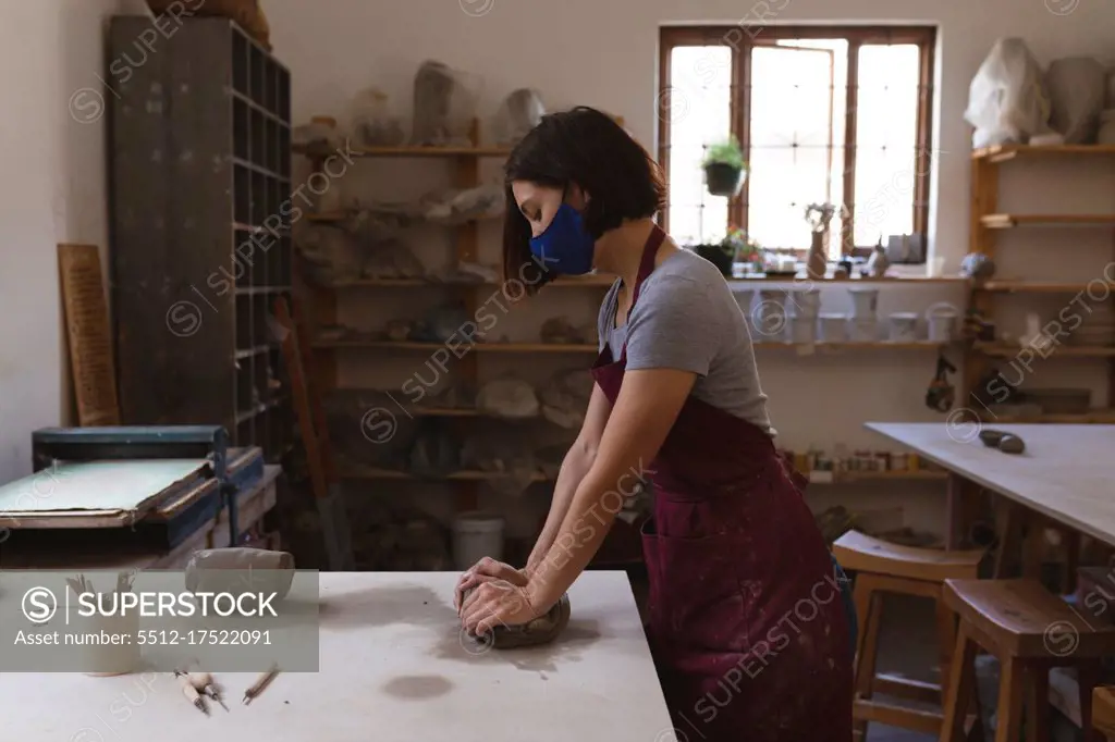 Caucasian female potter in face mask working in pottery studio. wearing apron, working at a working table. small creative business during covid 19 coronavirus pandemic.