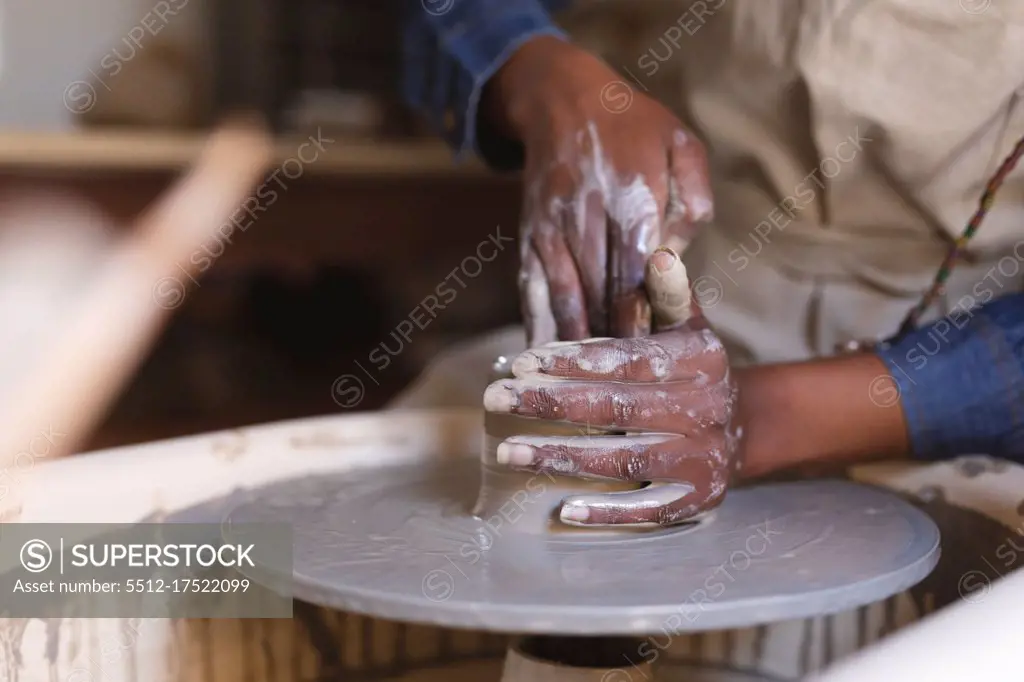 Mixed race female potter in face mask working in pottery studio. wearing apron, working at a potters wheel. small creative business during covid 19 coronavirus pandemic.
