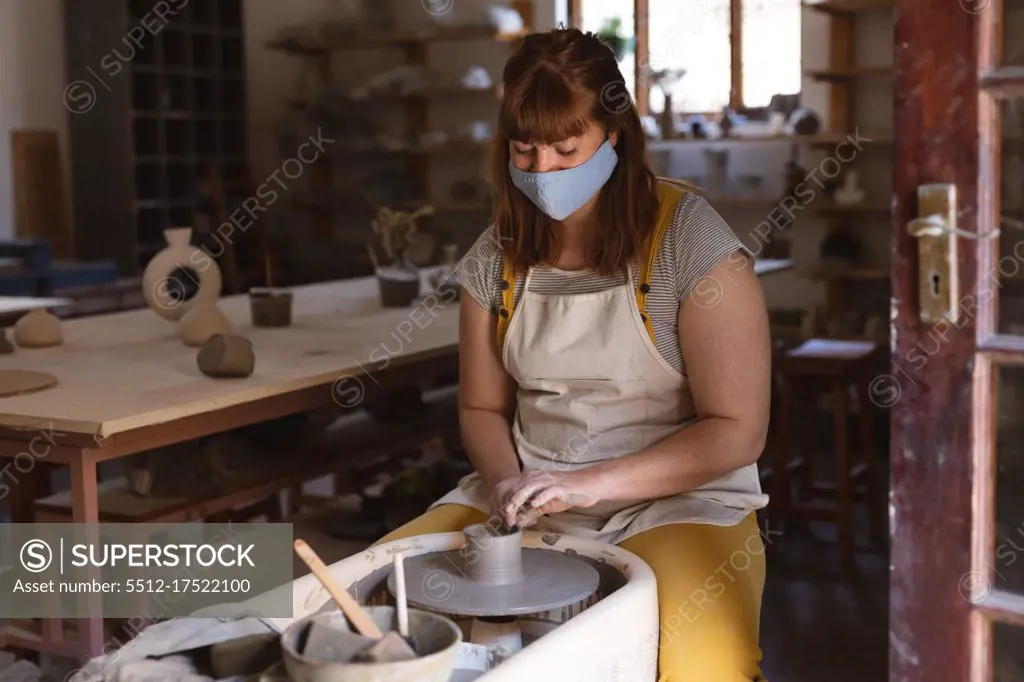 Caucasian female potter in face mask working in pottery studio. wearing apron, working at a potters wheel. small creative business during covid 19 coronavirus pandemic.