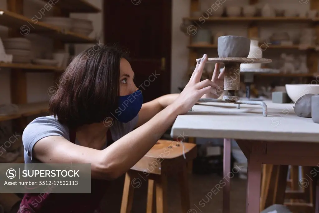 Caucasian female potter in face mask working in pottery studio. wearing apron, working at a potters wheel. small creative business during covid 19 coronavirus pandemic.