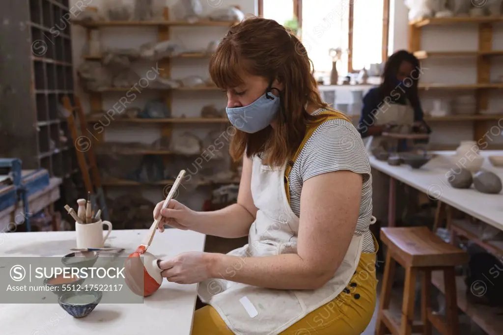 Caucasian female potter in face mask working in pottery studio. wearing apron, working at a working table, painting a plate. small creative business during covid 19 coronavirus pandemic.