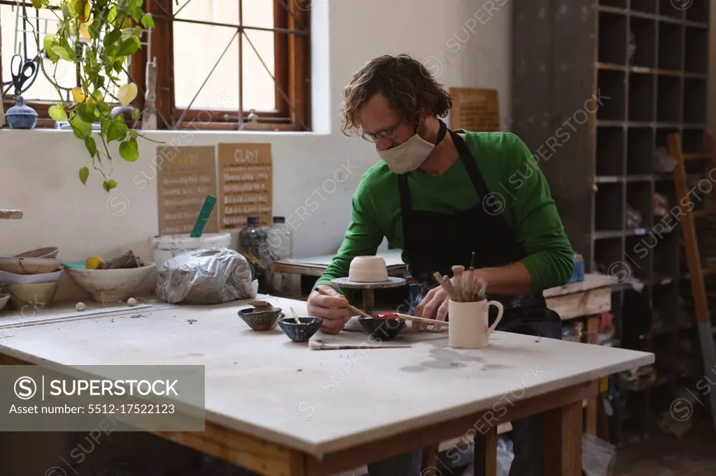 Caucasian male potter in face mask working in pottery studio. wearing apron, working at a potters wheel, painting a bowl. small creative business during covid 19 coronavirus pandemic.
