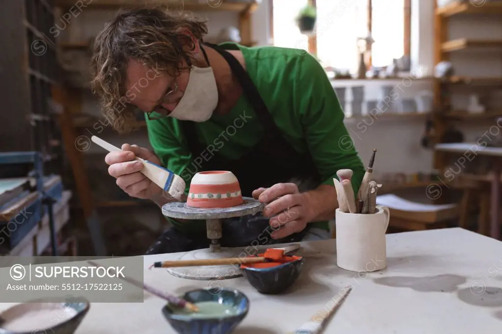Caucasian male potter in face mask working in pottery studio. wearing apron, working at a potters wheel, painting bowl. small creative business during covid 19 coronavirus pandemic.
