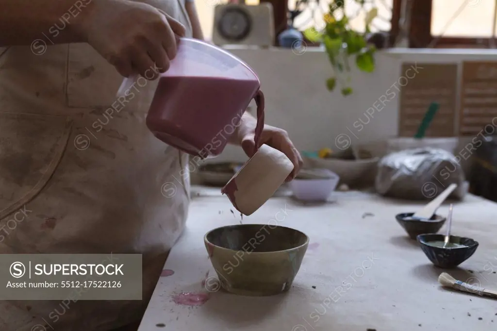 Caucasian female potter in face mask working in pottery studio. wearing apron, working at a potters wheel, painting a bowl. small creative business during covid 19 coronavirus pandemic.