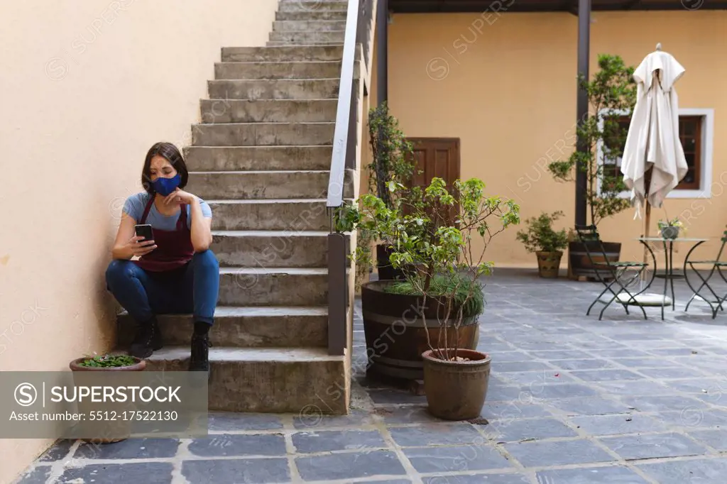 Caucasian female potter sitting on stairs outside pottery studio. wearing apron, using smartphone. small creative business during covid 19 coronavirus pandemic.