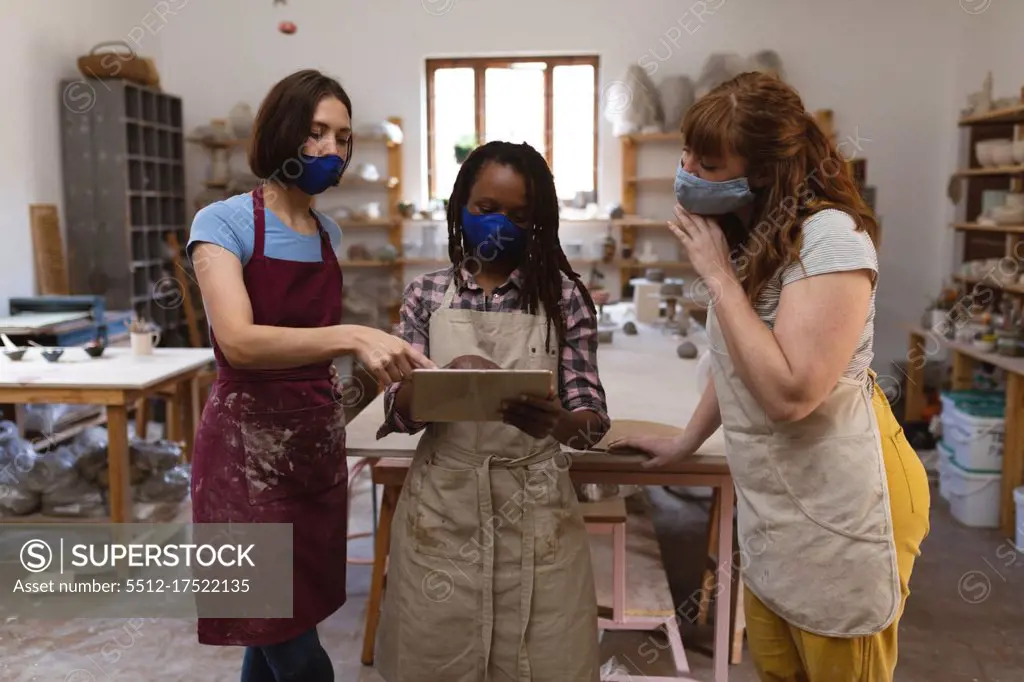 Two caucasian and one mixed race female potters in face mask working in pottery studio. wearing aprons, looking at digital tablet. small creative business during covid 19 coronavirus pandemic.