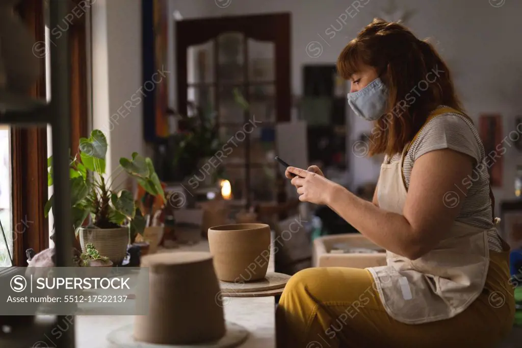 Caucasian female potter in face mask working in pottery studio. wearing apron, taking a photo of a bowl. small creative business during covid 19 coronavirus pandemic.