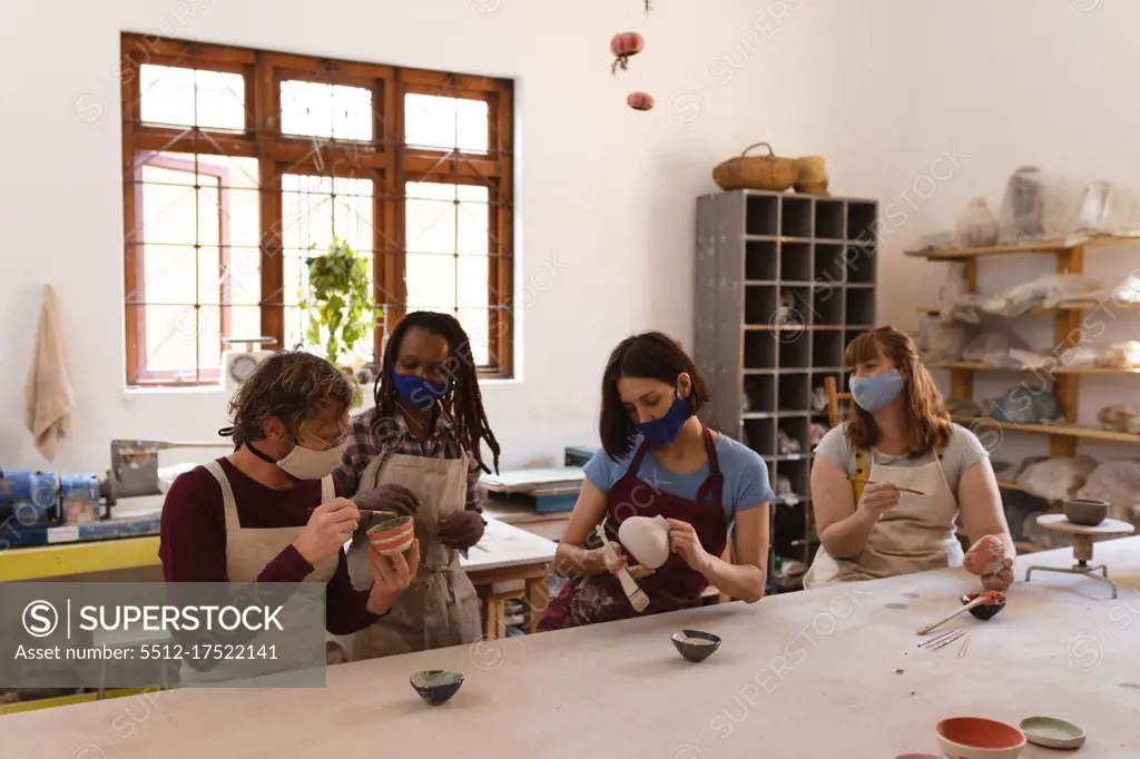 Multi-ethnic group of potters in face masks working in pottery studio. wearing aprons, painting plates. small creative business during covid 19 coronavirus pandemic.