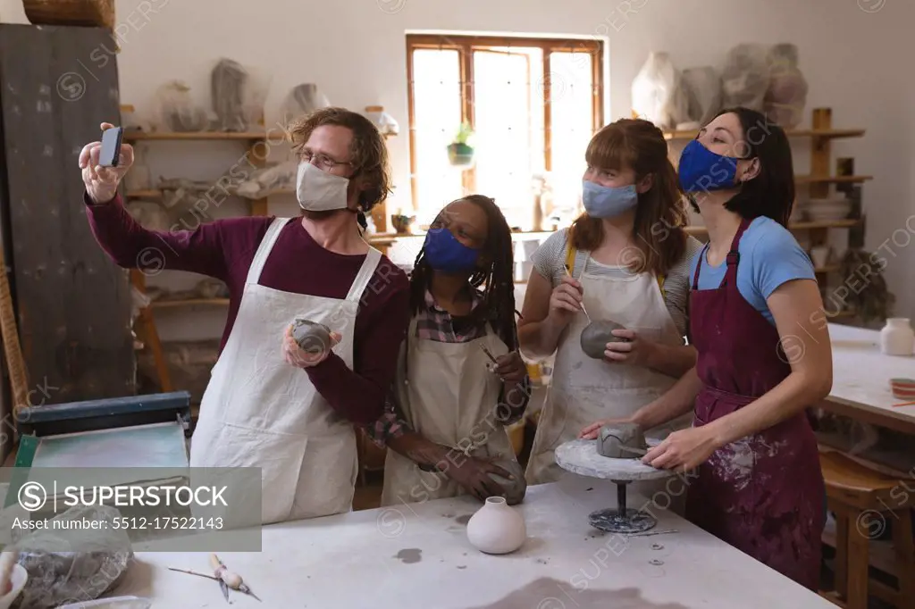 Multi-ethnic group of potters in face masks working in pottery studio. wearing aprons, painting plates, taking a selfie together. small creative business during covid 19 coronavirus pandemic.