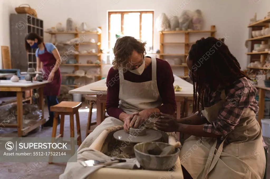 Caucasian male and mixed race female potters in face masks working in pottery studio. wearing aprons, working at a potters wheel. small creative business during covid 19 coronavirus pandemic.