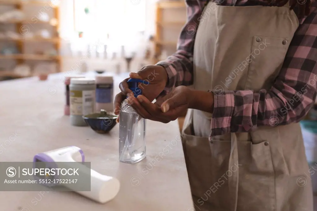 Mixed race female potter in face mask working in pottery studio. wearing apron, disinfecting her hands. small creative business during covid 19 coronavirus pandemic.