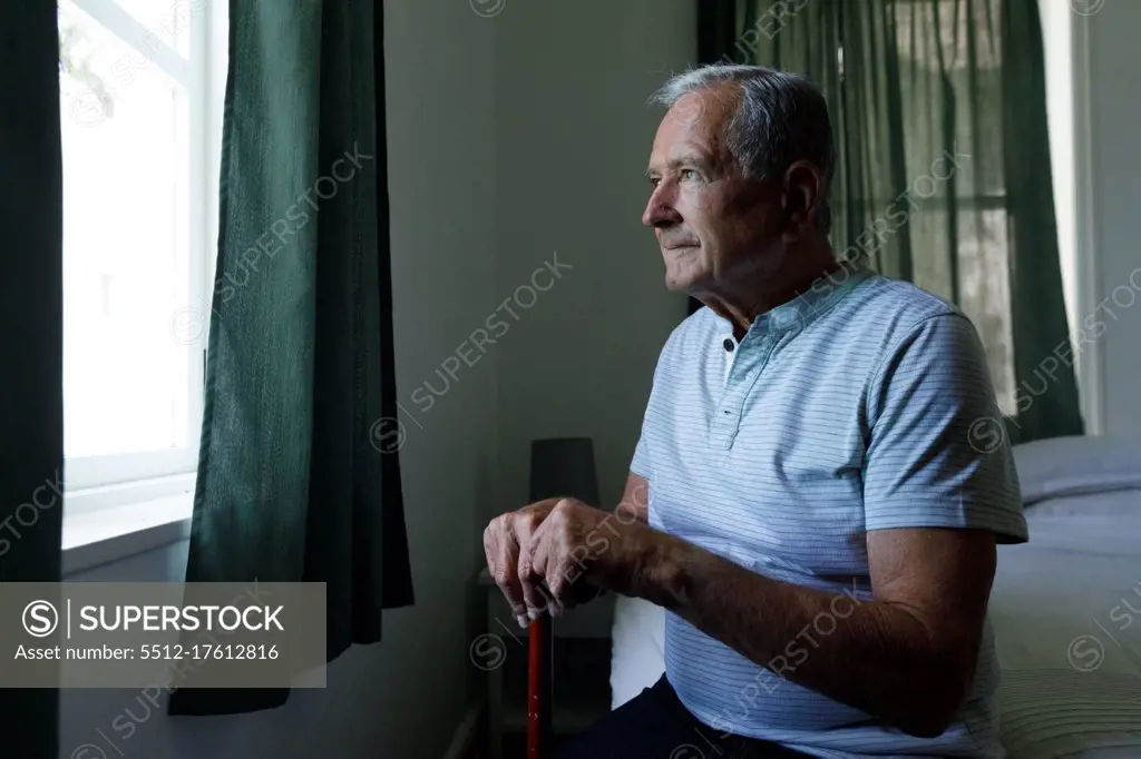 Senior caucasian man holding walking stick looking out of window at home. social distancing quarantine lockdown during coronavirus pandemic