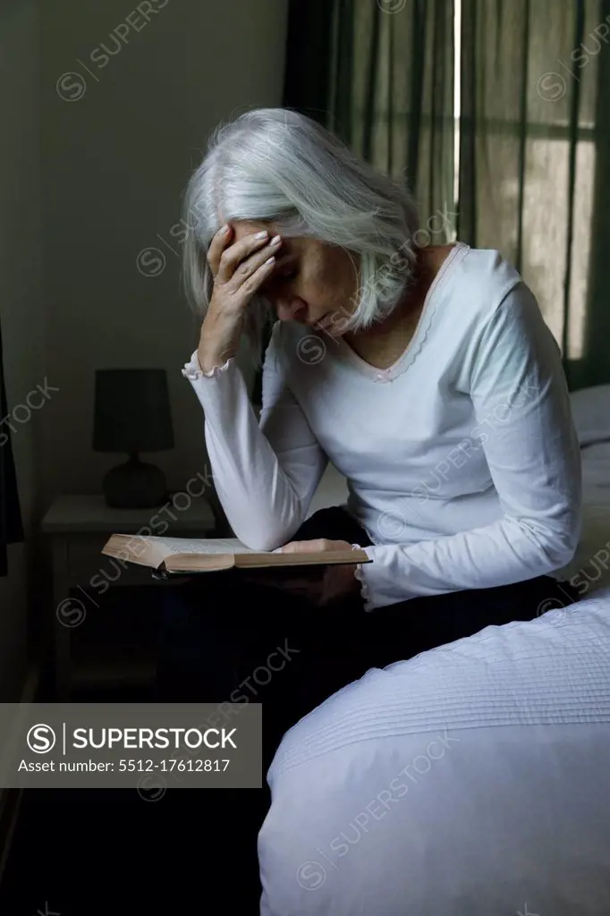 Stressed senior caucasian woman reading book while sitting on bed at home. social distancing quarantine lockdown during coronavirus pandemic