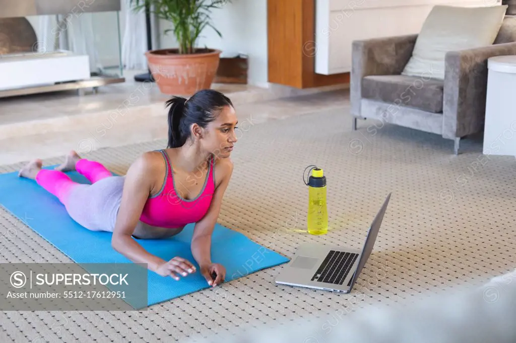 Mixed race woman practising yoga at home. using her laptop. self isolation during covid 19 coronavirus pandemic.