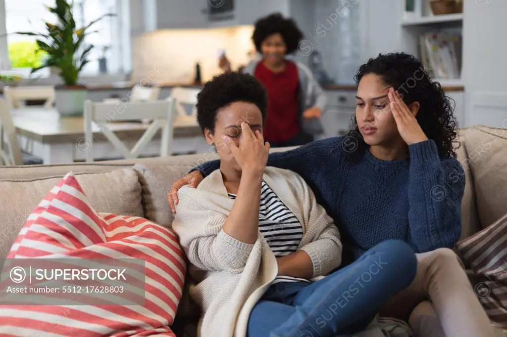 Upset mixed race lesbian sitting on couch. daughte rin the background. self isolation quality family time at home together during coronavirus covid 19 pandemic.