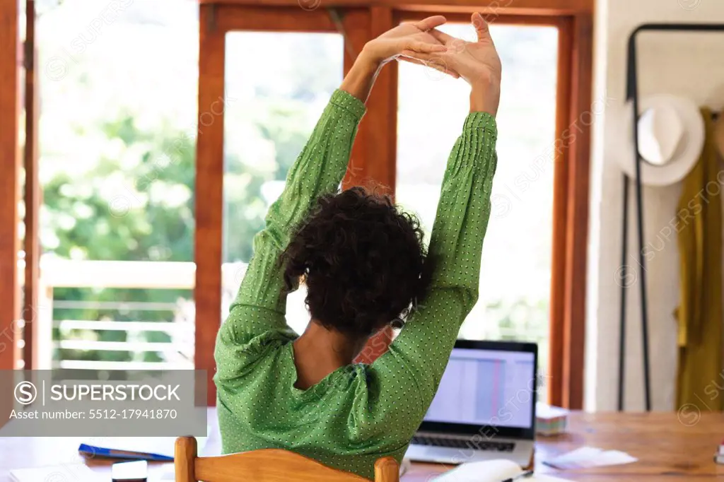 Rear view of caucasian woman sitting at table using laptop computer stretching arms in the air. staying at home in self isolation during quarantine lockdown.