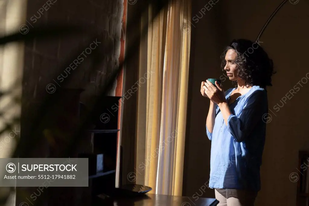 Thoughtful caucasian woman holding green mug looking out of window at home. Staying at home in self isolation during quarantine lockdown.