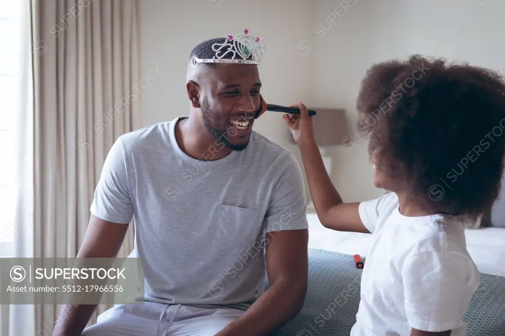 African american man wearing tiara having makeup put on by his daughter. staying at home in self isolation during quarantine lockdown.