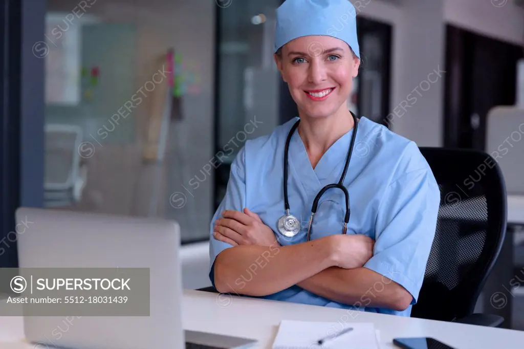 Portrait of smiling caucasian female doctor sitting at desk wearing scrubs using laptop computer. medical professional at work.