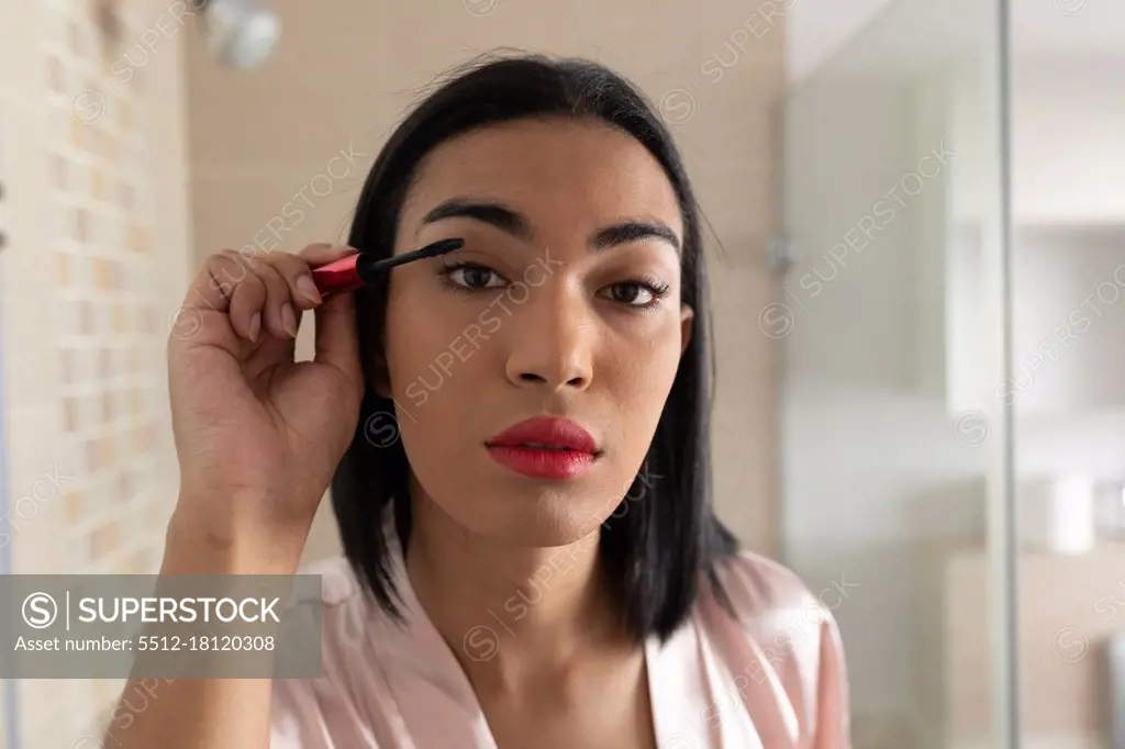 Portrait of mixed race transgender woman reflected in bathroom mirror putting on mascara. staying at home in isolation during quarantine lockdown.