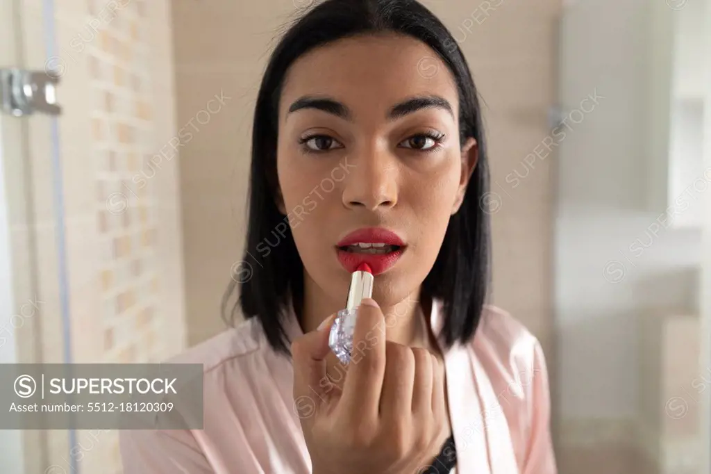 Portrait of mixed race transgender woman reflected in bathroom mirror putting on lipstick. staying at home in isolation during quarantine lockdown.