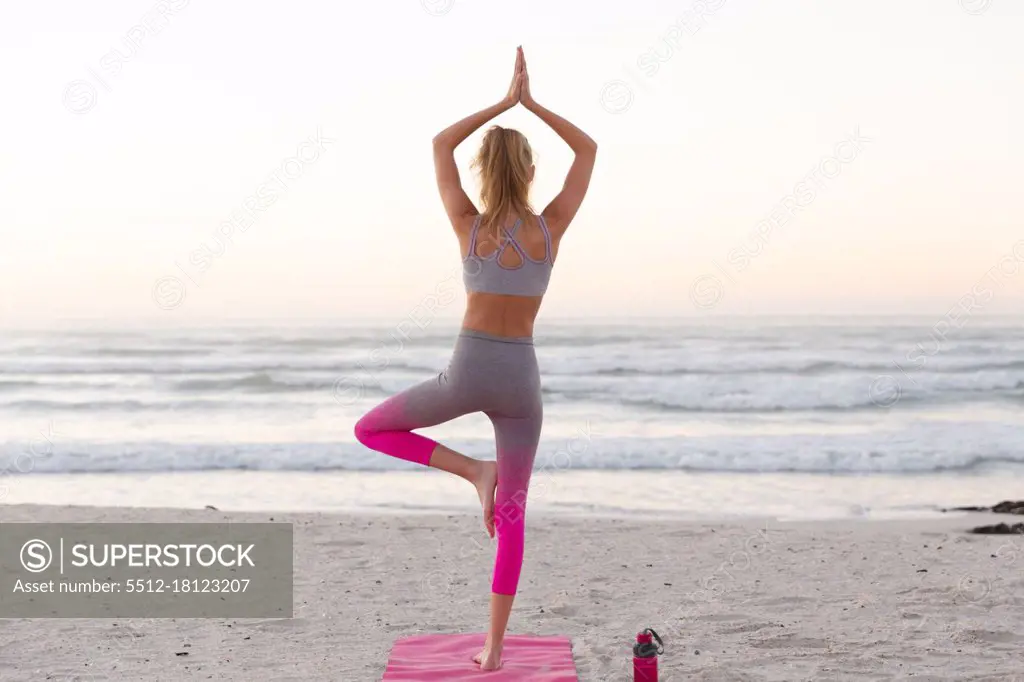 Rear view of of caucasian woman meditating and practicing yoga while standing on yoga mat at the beach. fitness yoga and healthy lifestyle concept