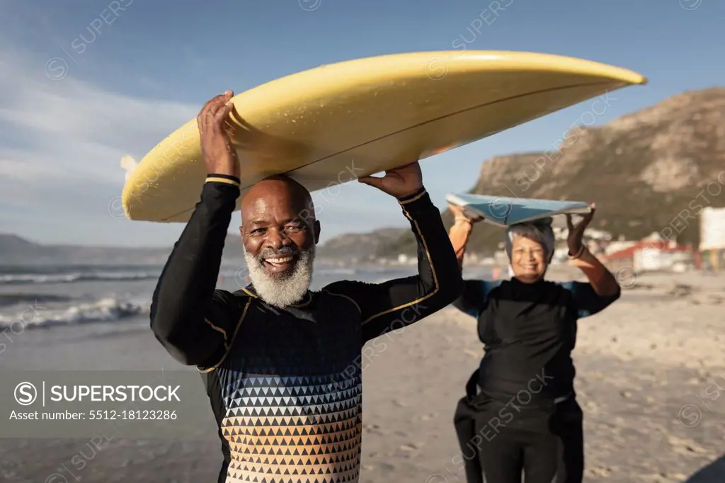 Portrait of senior african american couple carrying surf board on their head on the beach. travel vacation retirement lifestyle concept