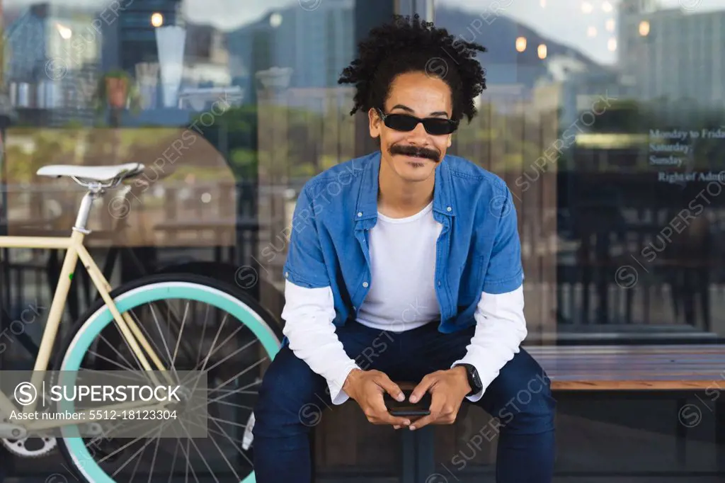 Portrait of happy mixed race man with moustache sitting on bench in street using smartphone. digital nomad, out and about in the city.