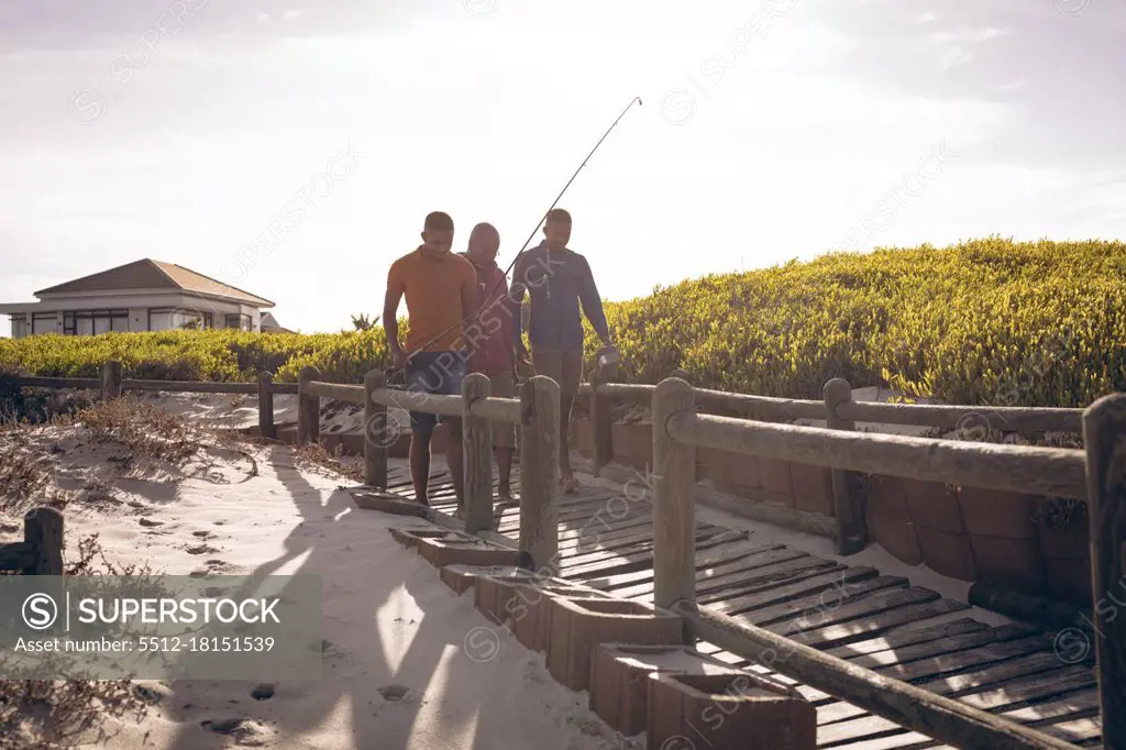 African american father and his two sons with fishing rods walking together on the bridge. summer beach holiday and leisure concept.