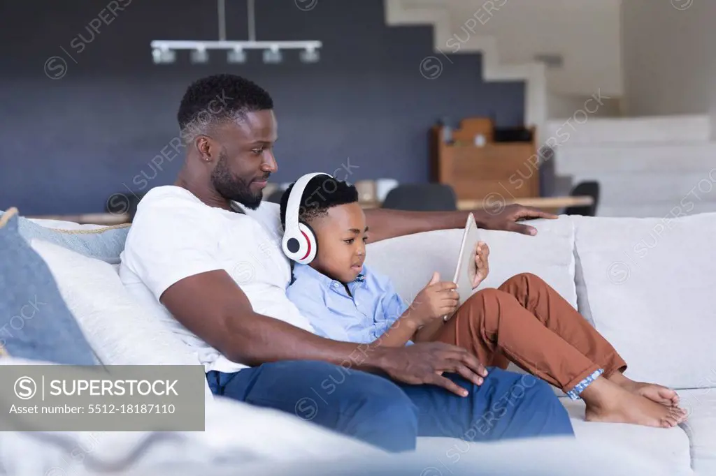 African american father and son sitting on sofa, using tablet and smiling. at home in isolation during quarantine lockdown.