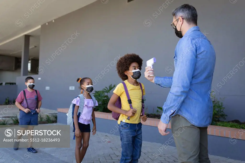 Caucasian male teacher measuring temperature of students standing in a queue at elementary school. hygiene and social distancing at school during covid 19 pandemic