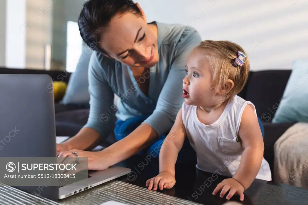 Smiling caucasian mother looking at her baby using laptop while working from home. motherhood, love and baby care concept