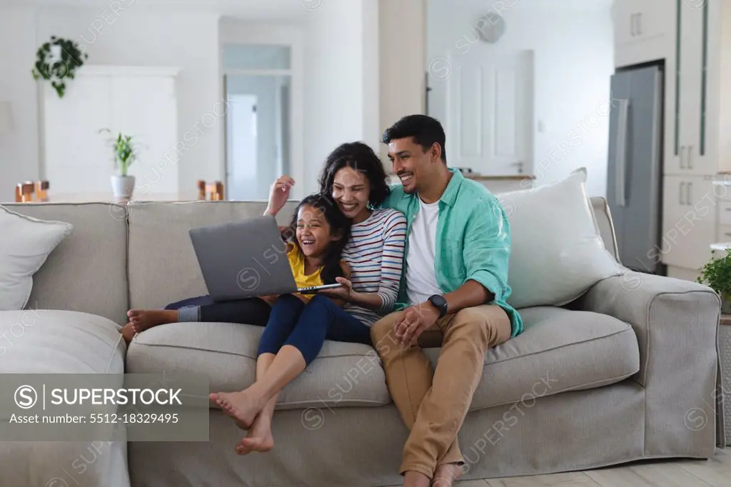 Smiling hispanic mother, father and daughter sitting on couch using laptop together. family spending time together at home.
